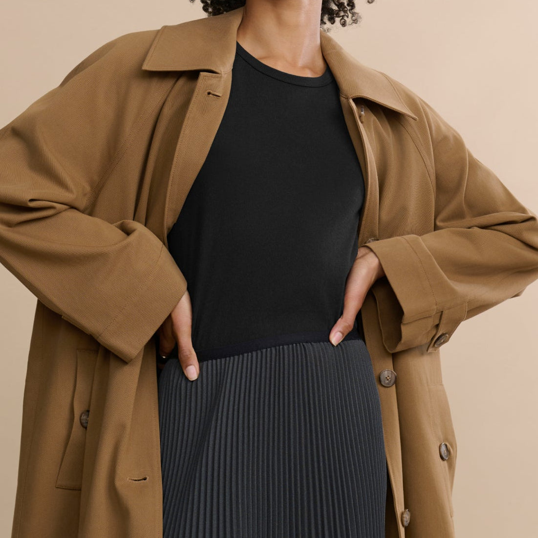 Woman wearing a brown coat over a black top and pleated skirt on a beige background