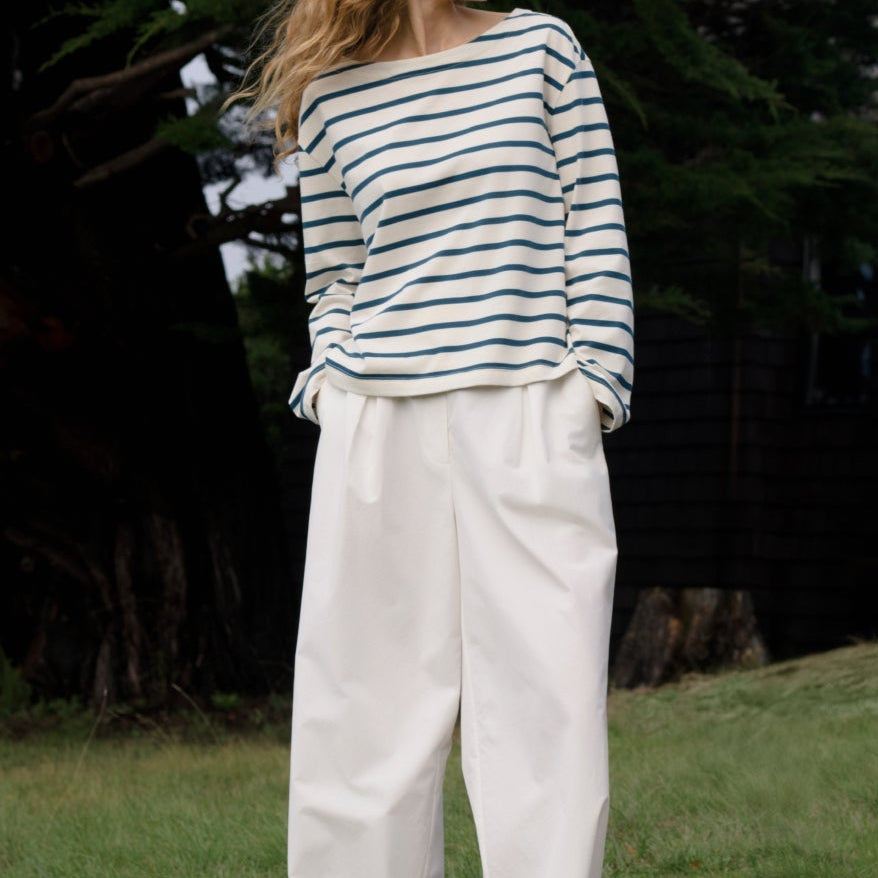 Woman wearing a striped shirt and white pants standing in a grassy area with trees and a building in the background.