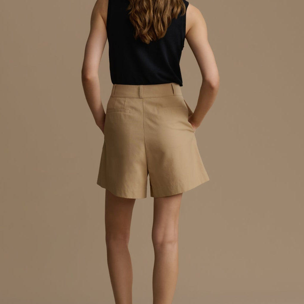 Woman wearing a black sleeveless top and beige shorts against a brown background