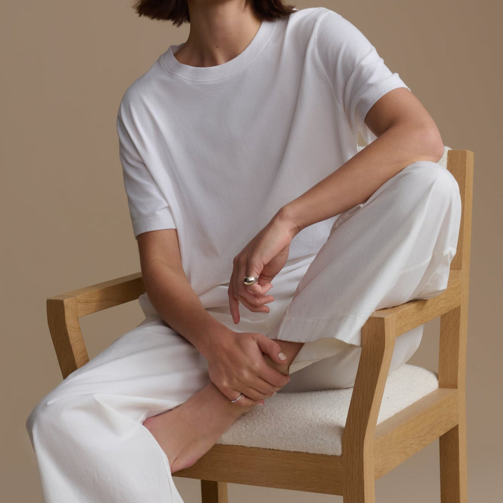 Woman sitting on a wooden chair wearing a white outfit against a beige background
