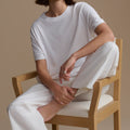 Woman sitting on a wooden chair wearing a white outfit against a beige background