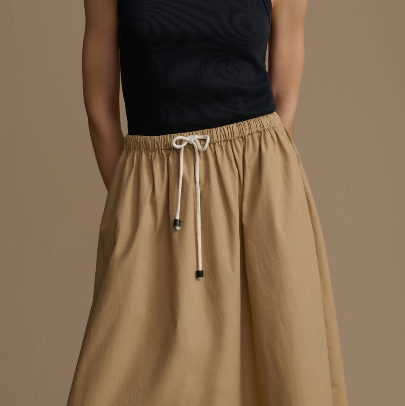 Person wearing a black sleeveless top and tan drawstring pants on a brown background