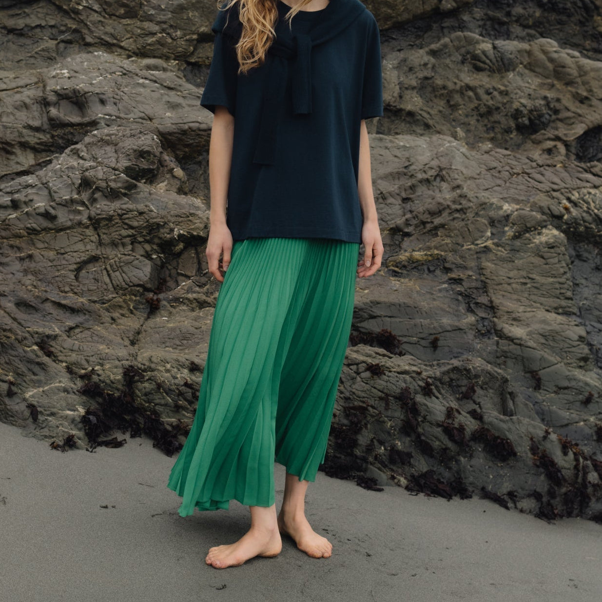 Woman in a black top and green pants standing on a rocky beach.