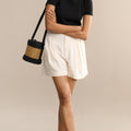 Woman wearing a black top and white shorts, holding a black and beige handbag against a beige background