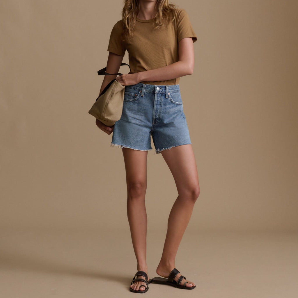 Woman wearing a brown t-shirt and blue denim shorts holding a beige handbag against a beige background